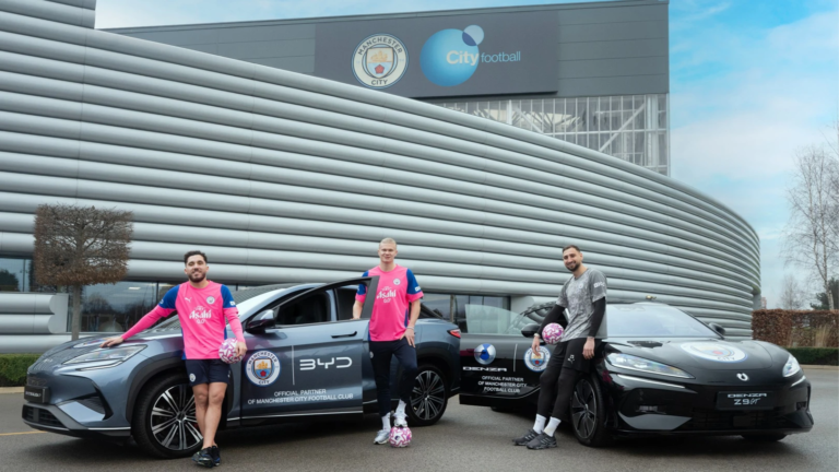 BYD branding on Manchester City's training kit and electric vehicles parked at the City Football Academy.